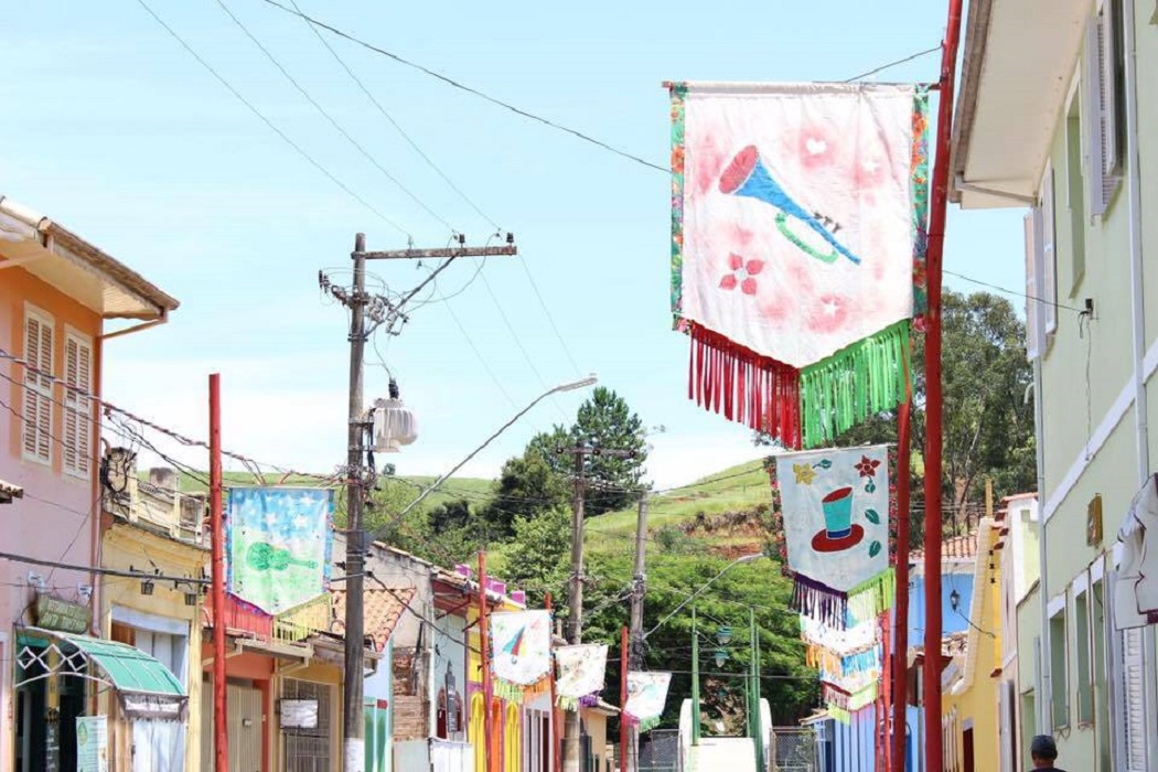 Foto mostra decoração de Carnaval em São Luiz do Paraitinga, com céu azul, cabos passando, montanhas verdes ao fundo e casas coloridas.