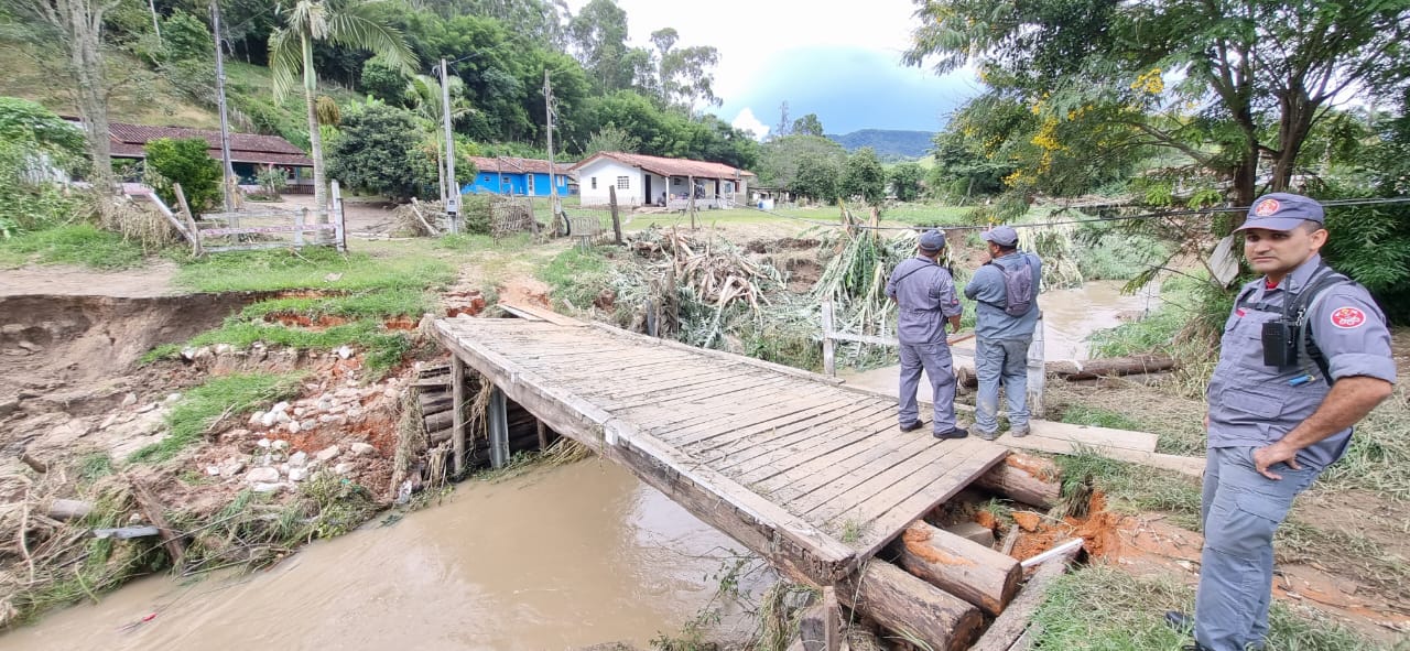 Bombeiros auxiliam nas chuvas em São Luiz do Paraitinga; foto mostra lago com uma ponte em cima, árvores em volta, céu azul e uma casa ao fundo.