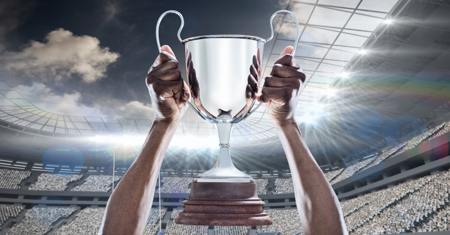 Mãos segurando uma taça, com um estádio ao fundo; foto ilustra matéria sobre o mundo da bola em fevereiro.