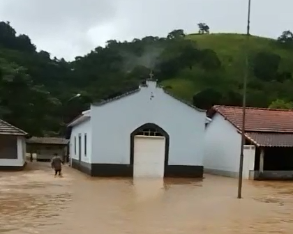 Igreja da Pedra Branca, em Cunha, fica alagada após chuvas. Foto mostra água na parte externa do local, com um homem andando e árvores ao fundo.