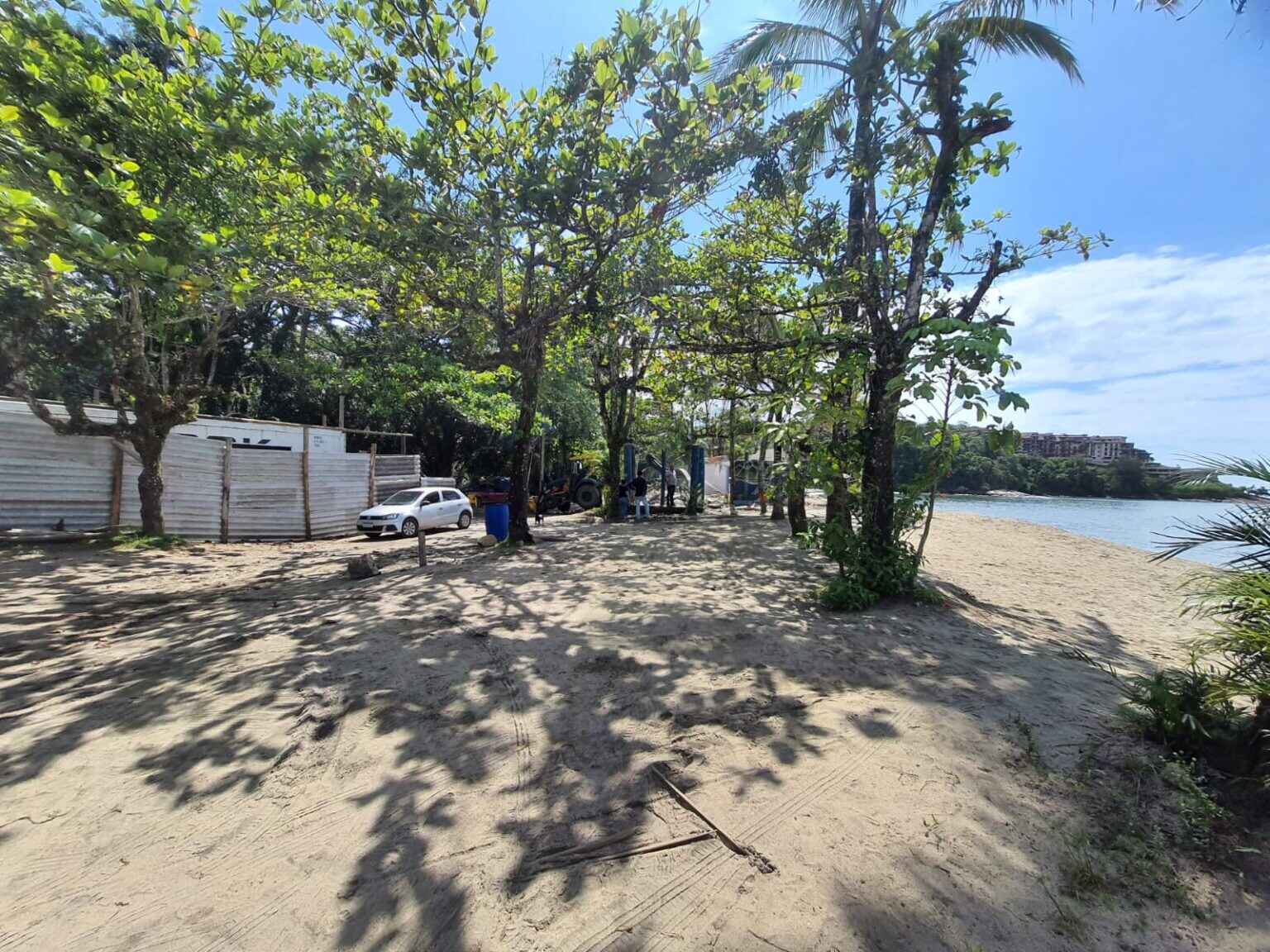 Praia da Mococa, em Caraguatatuba, com mar no lado direito, areia no lado esquerdo, árvores ao fundo e céu azul.