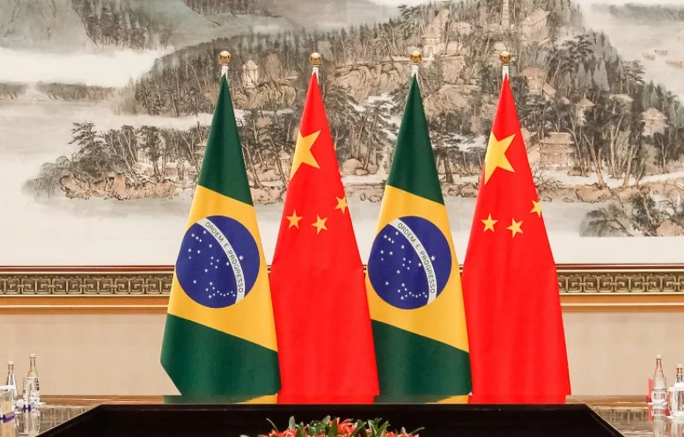 Bandeiras do Brasil e China, enfileiradas, com um quadro ao fundo, ilustrando matéria sobre acordo de São José dos Campos com a cidade Taishan; foto é de Beto Barata.