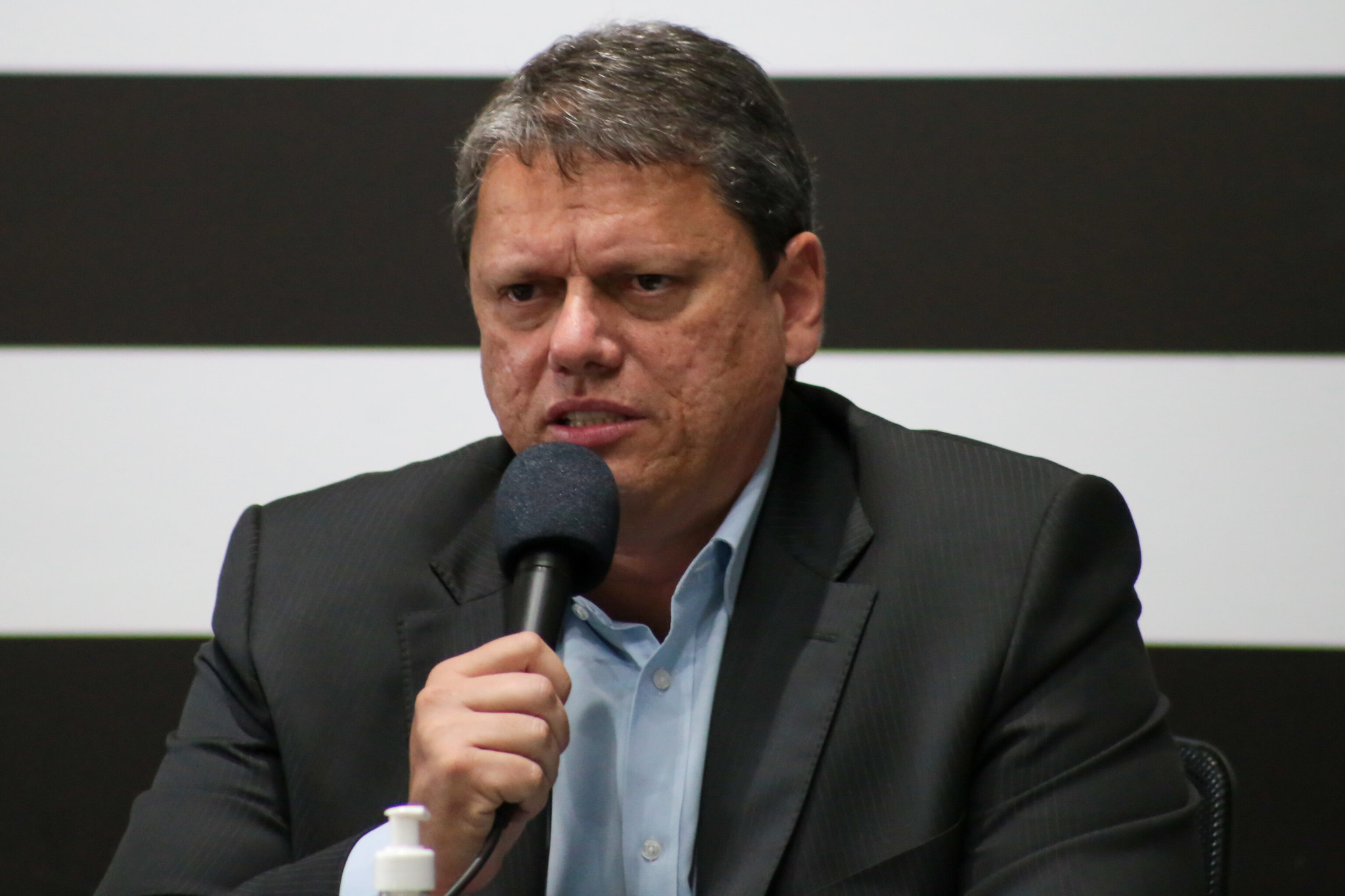 Tarcísio de Freitas com terno preto, camisa azul, falando ao microfone, em frente a uma parede preta e branca; foto ilustra matéria sobre o fim do passaporte vacinal em SP.