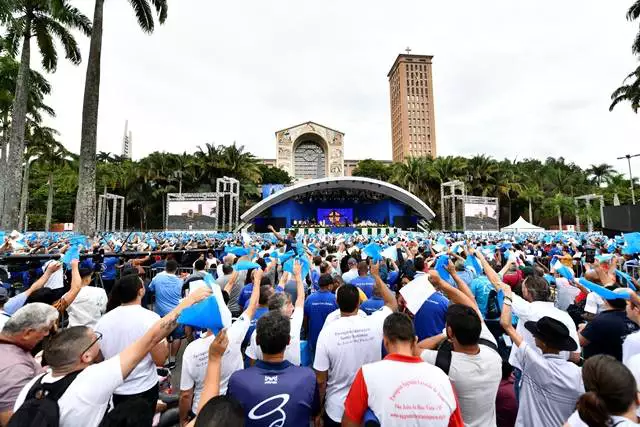 Terço dos Homens, em frente ao Santuário de Aparecida, com pessoas vestindo camisetas brancas, com detalhes em azul, céu azul e árvores nas laterais.