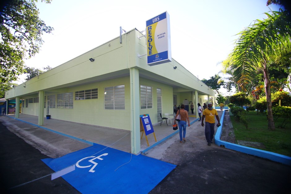 Fachada da UBS no Alto da Ponte, em São José dos Campos, com cores verdes, placa amarela, pessoas andando, árvores em volta e céu branco; foto ilustra matéria sobre agendamento de consultas do SUS pela Central 156.