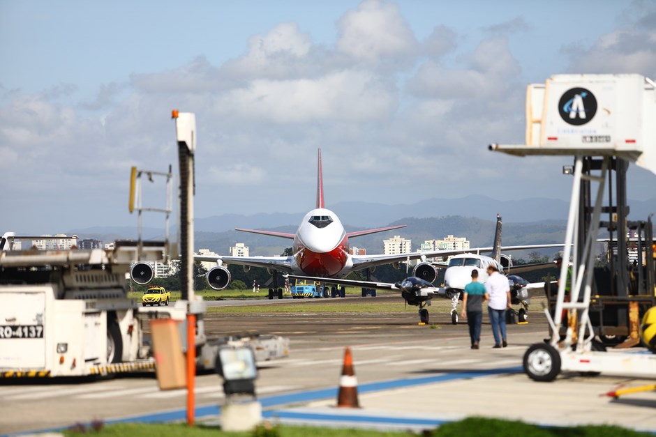 Aeroporto de SJC terá voos de passageiros ainda em 2023, afirmou Potiguara Campos; foto mostra um avião branco com detalhes vermelhos em uma pista, com céu azul e pessoas olhando, além de outros objetos ao redor.