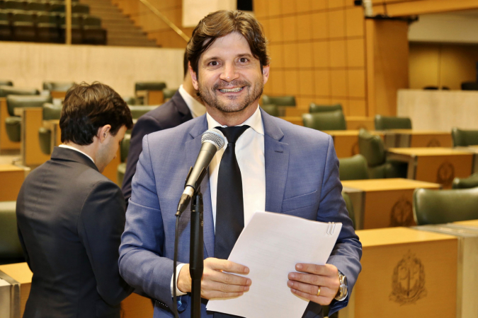 André do Prado com terno cinza, camisa branca, gravata preta, segurando um papel, falando no microfone e sorrindo; matéria ilustra sua posse como presidente da Alesp.