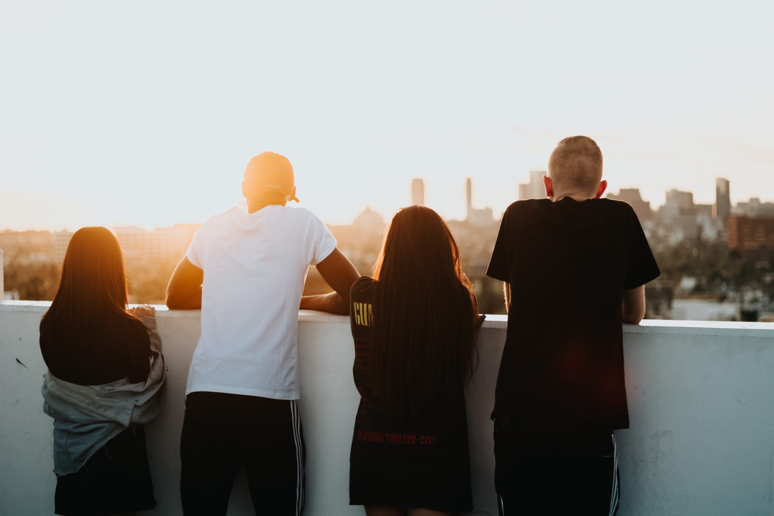 grupo de 4 jovens de costas olhando vista ao pôr do sol apoiados com os braços em muro branco.