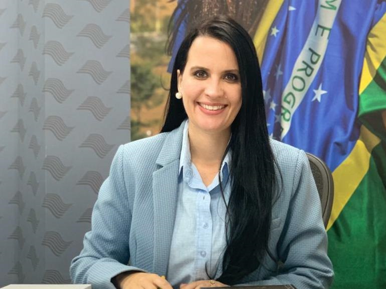 Retrato de Letícia Aguiar com terno e camisa azul, em frente a uma bandeira do Brasil, com cabelo preto e sorrindo; foto ilustra matéria sobre presidência do movimento Mulheres Progressistas.
