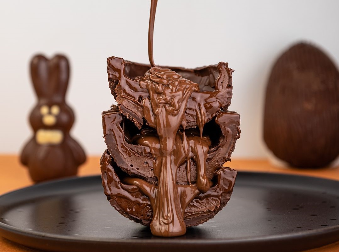 Foto de um chocolate da marca Lugano Chocolates, com uma calda sendo derramada, ao fundo uma parede branca e um coelho ao lado esquerdo.
