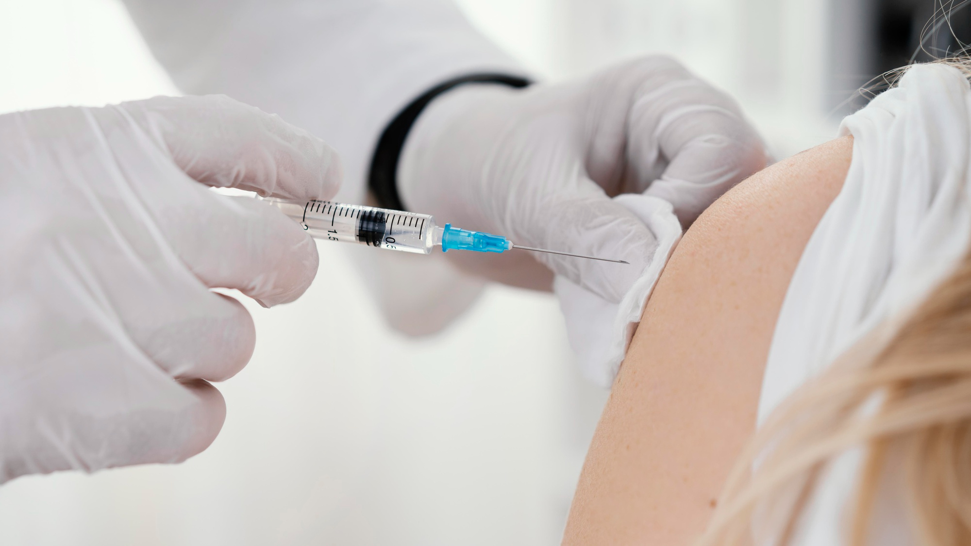 Profissional da saúde com uma luva branca vacinando um braço com um agulha transparente e detalhe em azul; foto ilustra matéria sobre novas recomendações da OMS sobre a dose de reforço contra a Covid-19.