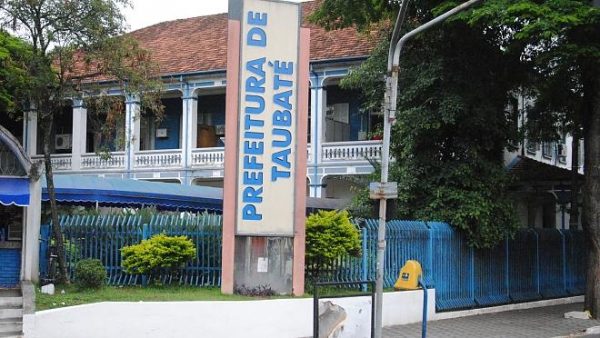 Fachada da Prefeitura de Taubaté, com um telhado em cima, uma placa no centro, árvores nas laterais e um portão azul; foto ilustra matéria sobre concurso público em Taubaté e vagas de emprego.
