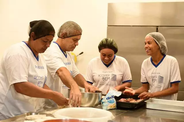 Alunos do curso profissionalizante em Aparecida, do Projeto Acolher Bem, do Santuário Nacional, com roupas brancas, touca, trabalhando em cima de uma mesa.