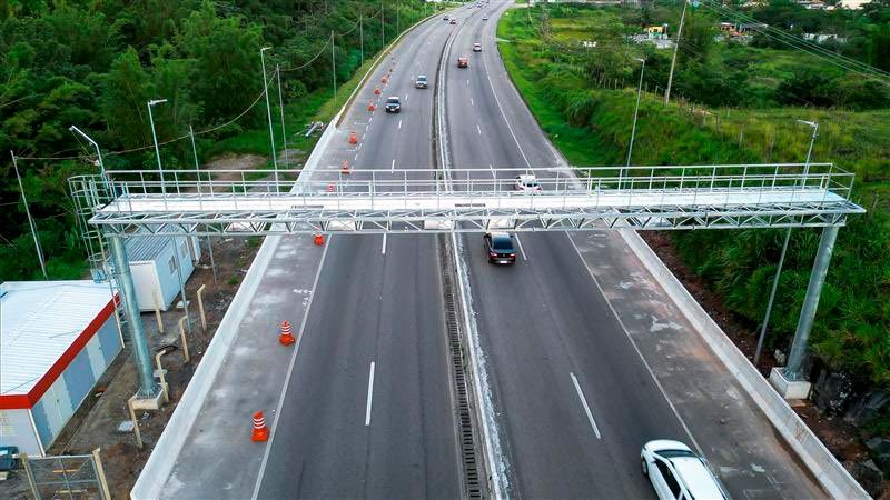 Vista aérea Rodovia Rio-Santos, com área verde ao lado, carros passando e um pedágio ao centro, ilustrando matéria sobre sistema free flow (autopagamento).