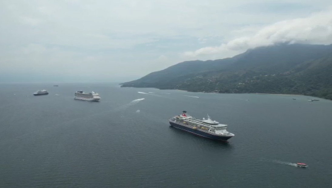 Foto aérea da temporada de navios em Ilhabela, com embarcações sob o mar, uma montanha ao fundo e céu azul; ilustrando matéria sobre os turistas.
