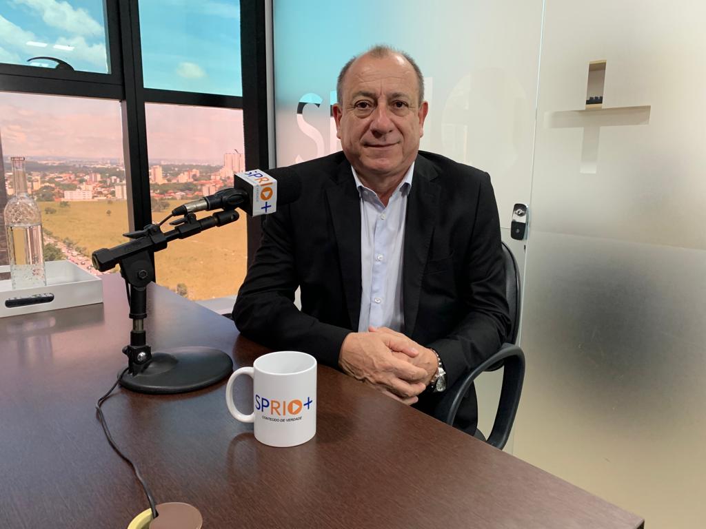Toni Sando, presidente da Unedestinos, sentado em uma cadeira preta, com os braços em uma mesa de madeira, com um microfone em um pedestal e uma caneca branca.