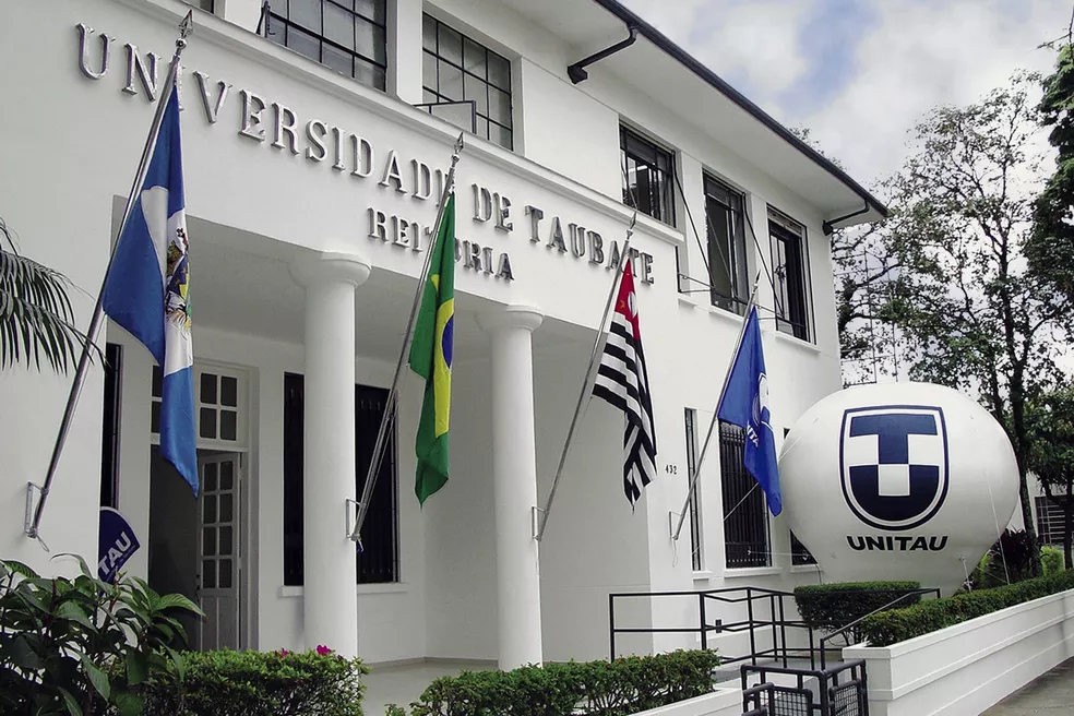 Fachada da Unitau, em Taubaté, com prédio branco, céu azul, bandeiras, logotipo da universidade e uma árvore; foto ilustra matéria sobre bolsas de estudo e proposta do Prouni municipal.