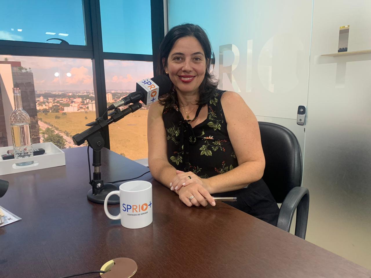 Vereadora Juliana Fraga, do PT de São José dos Campos, com roupa preta, sorrindo, com os braços apoiados em uma mesa de madeira e um vidro ao fundo.
