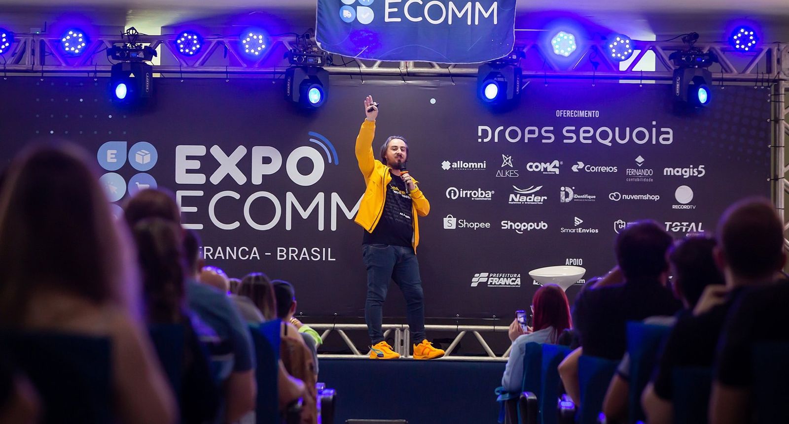 Paletrante durante uma edição da ExpoEcomm em frente a uma parede azul, com luzes e pessoas em volta; evento acontecerá em abril em São José dos Campos, no Parque Tecnológico.