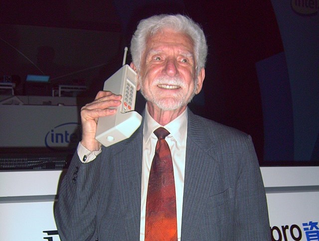 Martin Cooper com um celular apelidado de tijoão da Motorola, o DynaTAC 8000X, com terno preto, camisa branca e gravata vermelha.