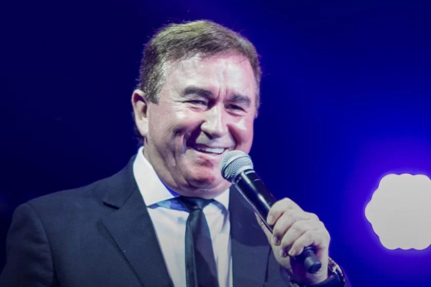 Foto ilustra matéria sobre o show de Amado Batista em SJC (São José dos Campos), que acontecerá no Palácio Sunset; retrato mostra o cantor com terno preto, camisa branca e gravata preta, enquanto sorri e fala em um microfone.