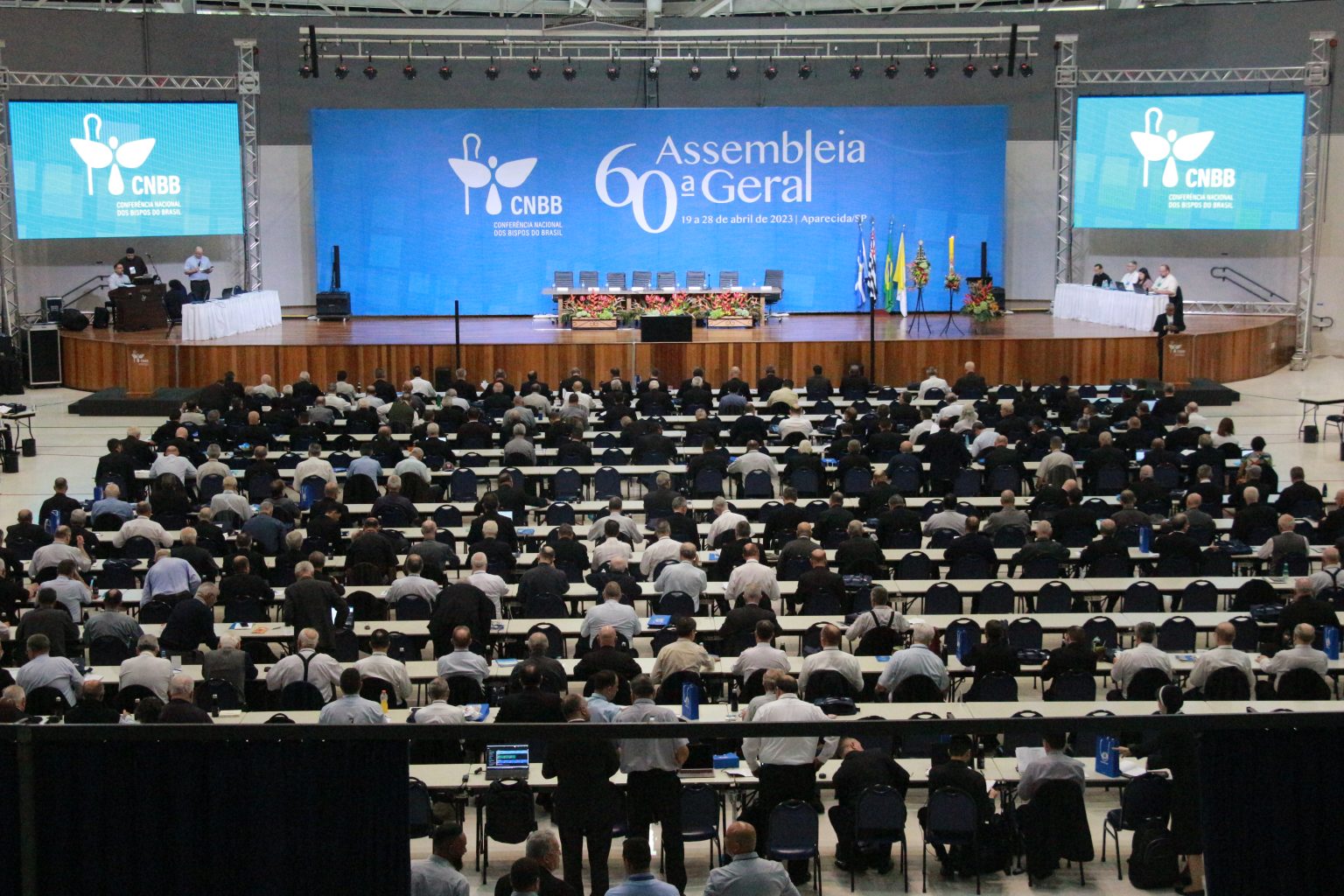 Foto da Assembleia da CNBB, realizada em Aparecida, com um telão azul ao centro, pessoas sentadas nas cadeiras e uma mesa de madeira.