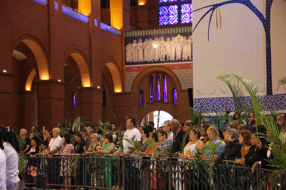 Santuário de Aparecida durante celebração do Domingo de Ramos 2023; foto mostra pessoas segurando ramos próximas a uma grade, com paredes marrons ao fundo e luzes.