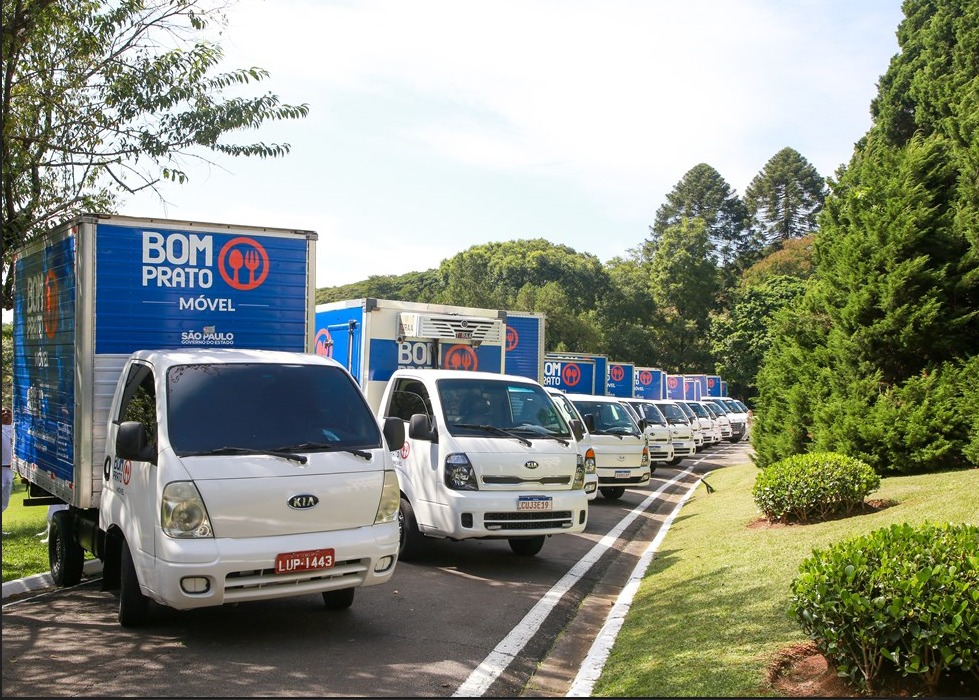 Veículos do Bom Prato Móvel Taubaté estacionados na rua, enfileirados, próximos a uma área verde, com um céu azul ao fundo.