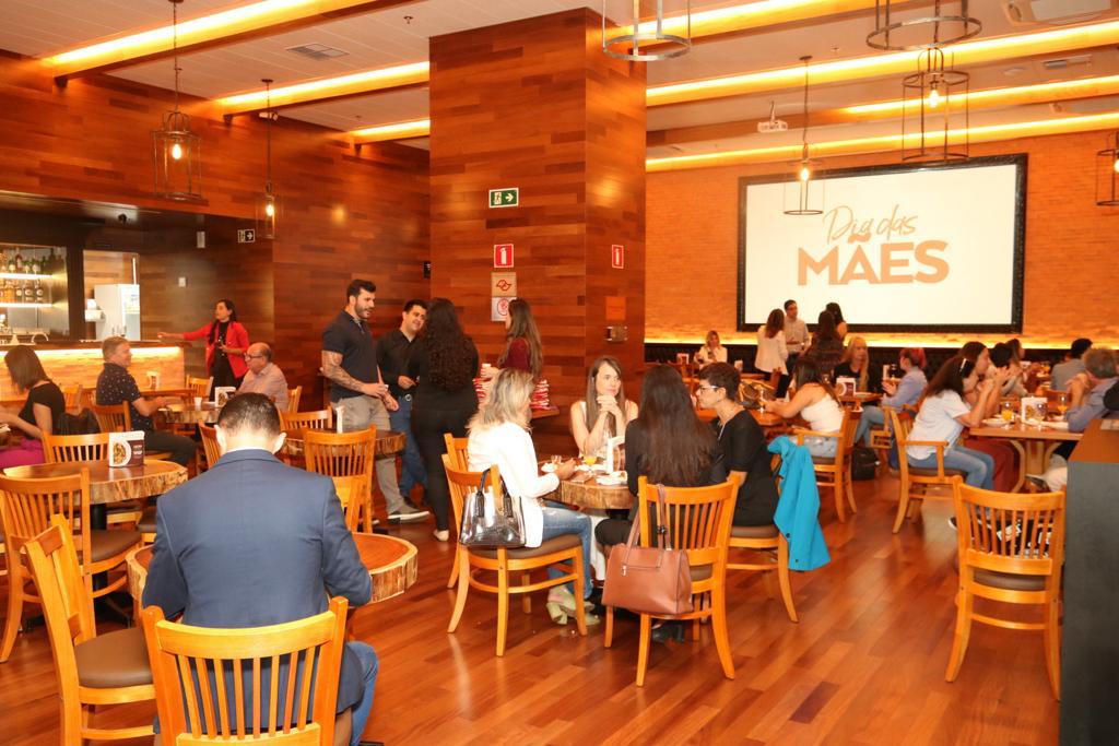 Jornalistas e influenciadores se reúnem no Madero do CenterVale Shopping, em São José dos Campos, para anúncio da campanha de Dia das Mães; foto tem decoração marrom com pessoas sentadas em cadeiras de madeira.