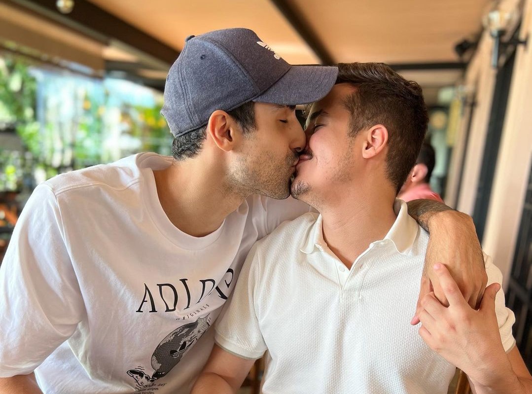 douglas souza, à esquerda, de camiseta branca e boné azul, beijando gabriel campos, seu noivo, à direita de camisa branca