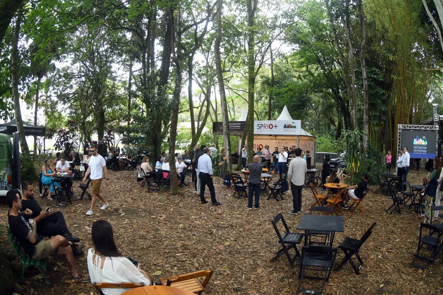 Área do festival +Gastronomia, realizado no Parque Vicentina Aranha, em São José dos Campos; foto mostra pessoas reunidas, em mesas, em meio à área verde com parvores e folhas no chão.