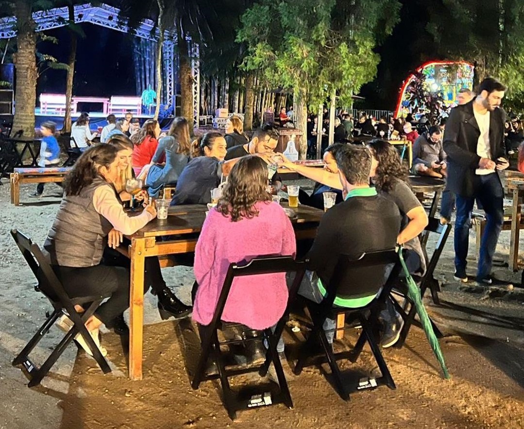 Pessoas com roupas coloridas brindando durante uma edição do Festival +Gastronomia, no Parque Vicentina Aranha, ao redor de uma mesa, com área verde ao fundo.