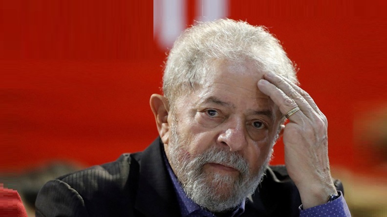 Retrato de Lula com a mão na testa, olhando para a câmera, usando um terno preto e uma camisa azul, em frente a uma parede vermelha; foto ilustra matéria sobre pesquisa que aponta o índice de aprovação e rejeição de seu governo.