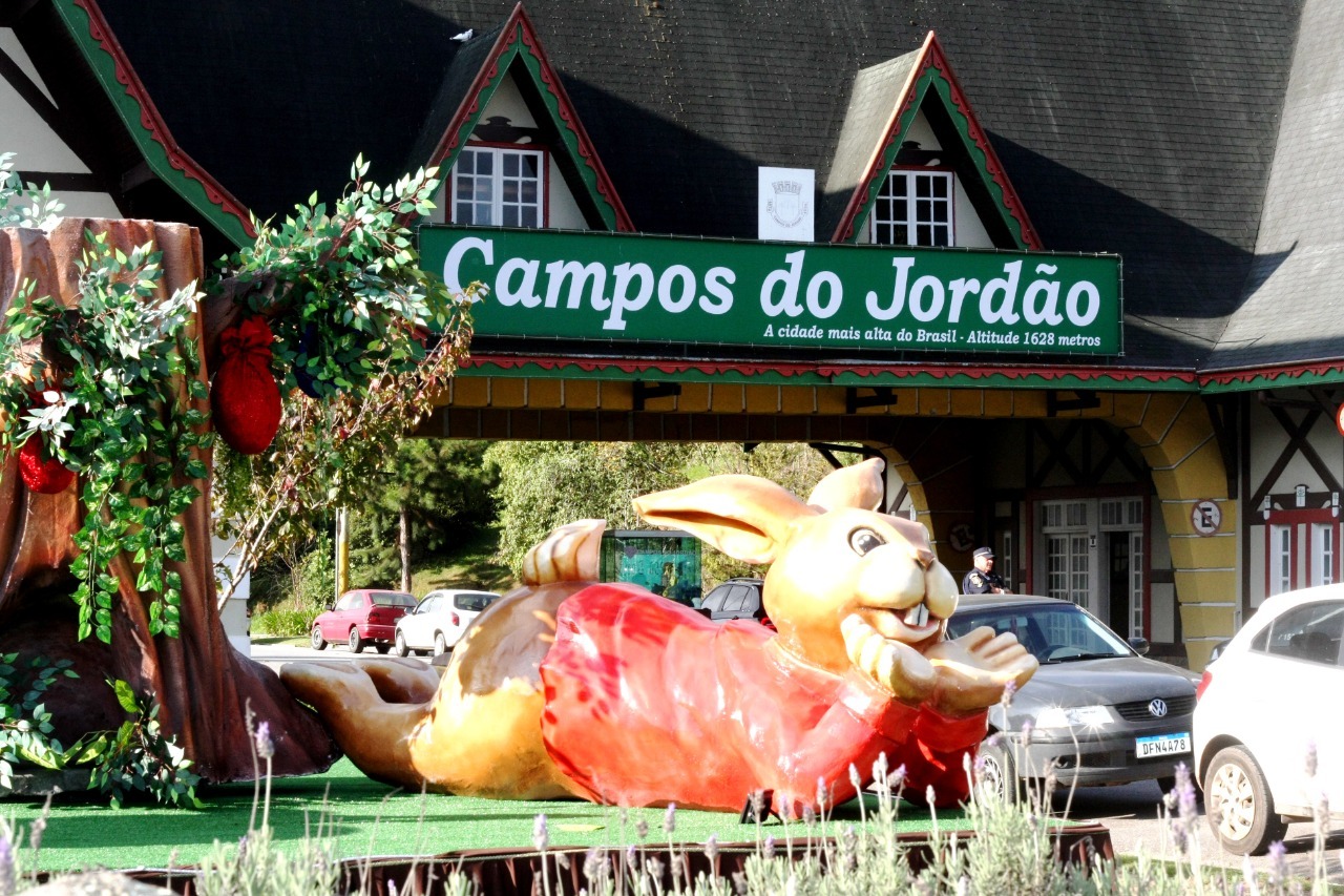 Portal de Campos do Jordão com decoração da Páscoa 2023, com um coelho gigante em frente, usando roupas vermelhas, enquanto carros passam ao fundo.