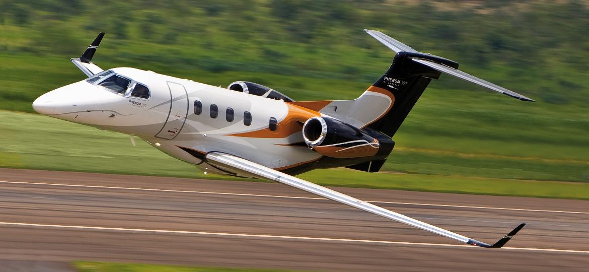Avião Phenom 300E, da Embraer, cuja sede é em São José dos Campos, voando próximo a uma pista, com árvores ao fundo; foto ilustra matéria sobre aeronave que apareceu na série O Agente Noturno, da Netflix.
