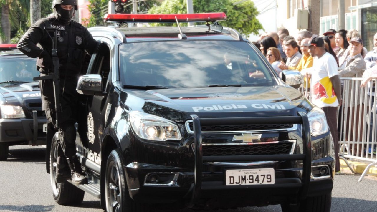 Integrantes da Polícia Civil do estado de São Paulo, em um carro preto, com pessoas ao redor observando.
