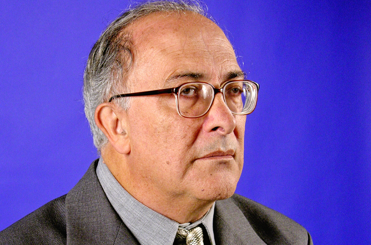 Professor Antônio Marmo, da Unitau, com um óculos preto, terno cinza, camisa e gravata, com um fundo azul.
