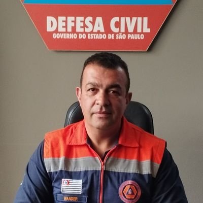 Retrato do secretário de Segurança de Campos do Jordão, Wander Vieira, com uma roupa laranja, cinza e azul, sentado em uma cadeira preta em frente a uma parede marrom.