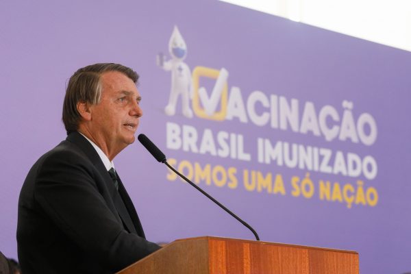 bolsonaro discursando em palanque com painel atrás que diz "