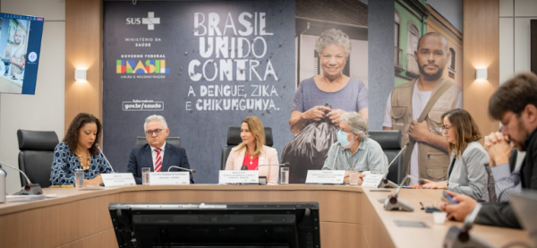Equipes do Governo Federal se reúnem para lançar campanha de combate à dengue no Brasil; foto mostra uma parede preta, com rostos na parede, uma mesa de madeira na frente, pessoas sentadas e uma tela de computador.