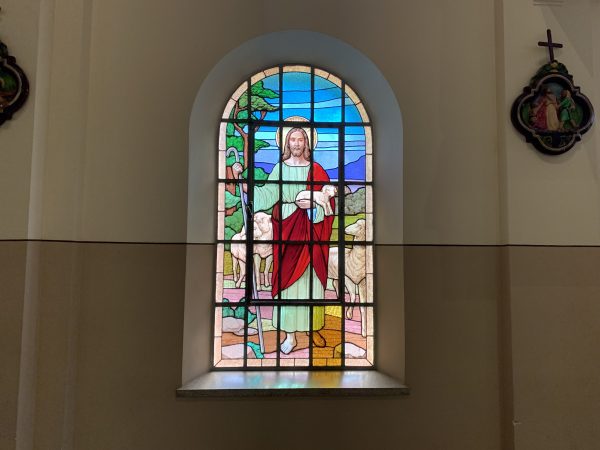 Imagem do Bom Pastor em vidro, com cores azul, vermelha e verde, em uma janela em uma parede marrom, com um quadro aos lados.