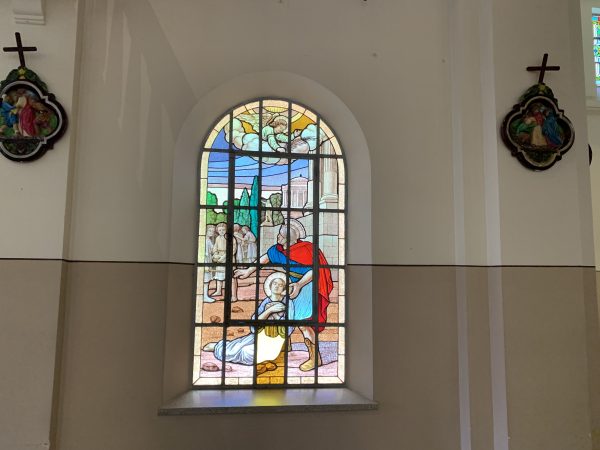 Imagem de São Tarcísio dentro da capela Sagrado Coração de Jesus, em vidros de uma janela, com parede marrom e bege; foto ilustra matéria sobre a história do local.