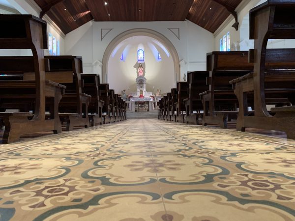 Piso de ladrilho hidráulico na capela, em SJC, com bancos de madeira, um altar no centro e uma imagem de Jesus ao fundo.