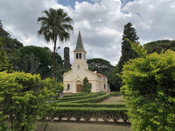 Fachada da capela do Vicentina Aranha, intitulada Sagrado Coração de Jesus, no parque na região central de São José dos Campos; foto mostra céu azul entre nuvens, área verde em volta do edifício, palmeira e um poste.