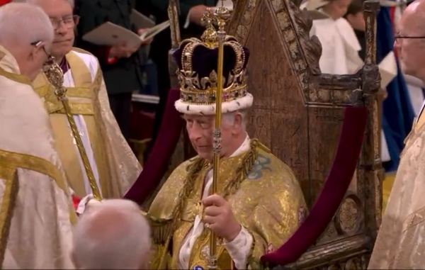 Rei Charless III, durante coroação no Reino Unido; na foto ele utiliza uma coroa e vestimentas douradas, enquanto está sentado em um trono de madeira, rodeado de pessoas.