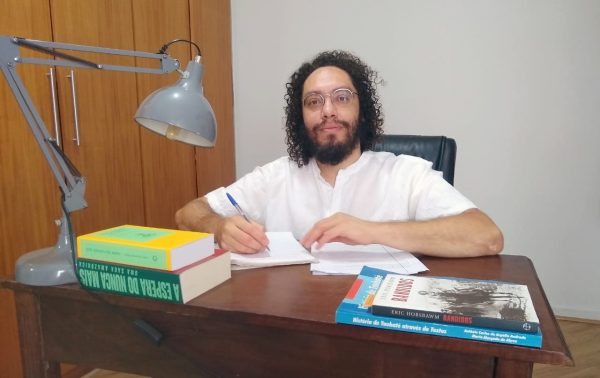 Rogério Guarapiran sentado, com os braços em uma mesa de madeira, vestindo uma camisa branca, sorrindo e olhando para a câmera; foto ilustra matéria sobre o projeto Bandoleiros de Taubaté.