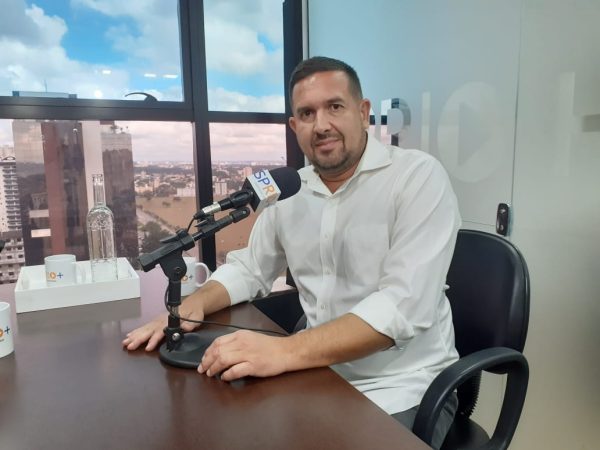 Alexandre Blanco, secretário de Projetos Especiais de São José dos Campos, sentado em uma cadeira preta, olhando para a câmera, com as mãos em uma mesa de madeira, sorrindo; foto ilustra matéria sobre a privatização do Mercado Municipal.