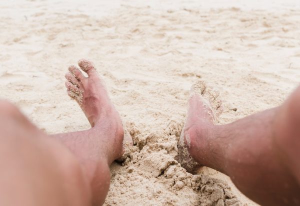 Pernas de um homem, com os pés na areia de uma praia; foto ilustra matéria sobre pesquisa da Unitau que revela risco de contaminação por parasitas em areias do Litoral Norte, como Caraguatatuba.