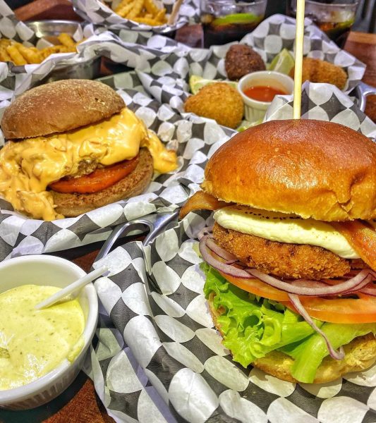 Lanches do Burguin, hamburgueria artesanal em São José dos Campos (SJC), em cima de um papel preto e branco, com molhos em volta e potes de batata frita ao fundo.