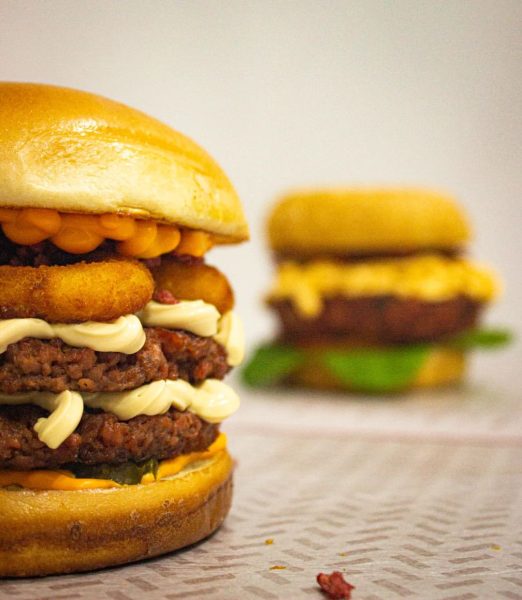 Uma opção de lanche no Caravegana, em São José dos Campos; foto mostra pão, hamburguer, molho e cheddar, em cima de uma mesa com toalha branca; ao fundo há outro lanche, mas desfocado.